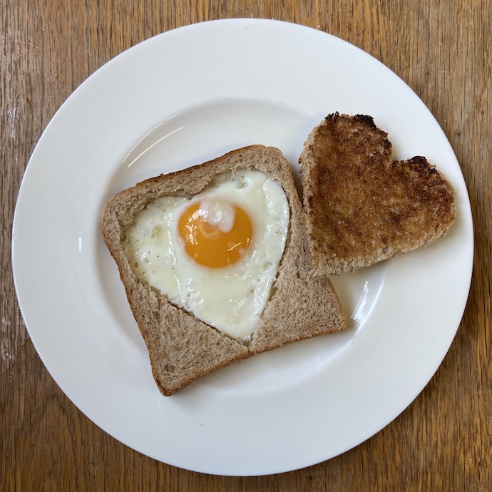 Picture of a heart shaped fried egg in a heart shaped hole in a piece of bread, with separate heart shaped piece of toast